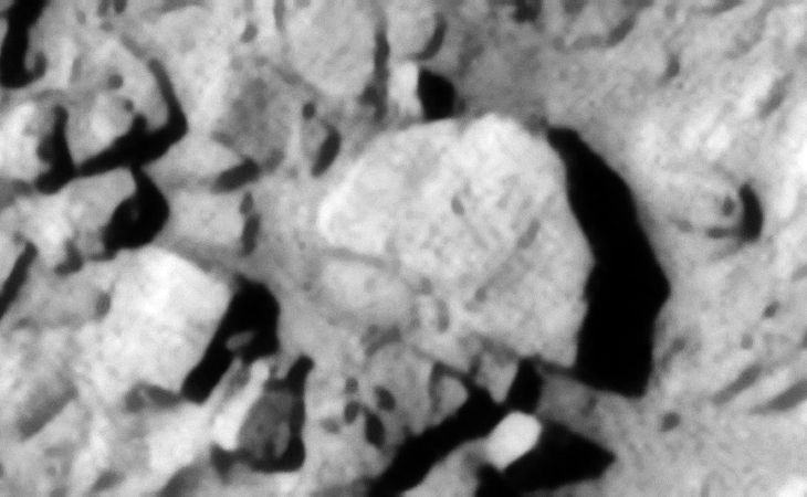 Semi-circular object located in the center of the image, it appears is may be on top stacked/layered metal plates. A smaller square megalithic block is seen to the left